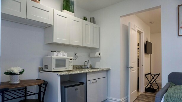 Une cuisine compacte avec des meubles blancs, un four à micro-ondes et un petit réfrigérateur. On y trouve une table à manger avec des chaises et une plante en pot.