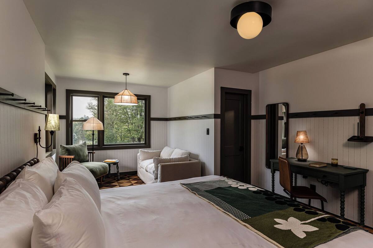 Ein Hotelzimmer mit einem großen Bett, einem Schreibtisch, einem Stuhl, einem kleinen Tisch und einem Fenster mit Blick auf Bäume.