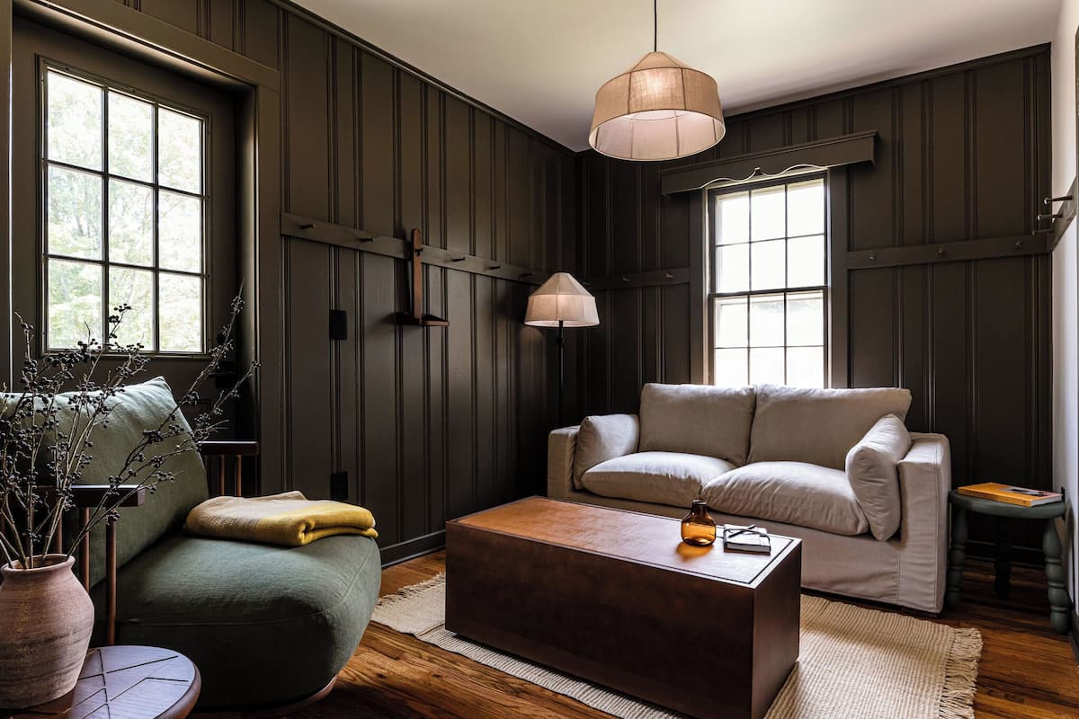 Ein Zimmer mit dunklen Holzwänden, einem grünen Sofa, einem beigen Sofa, einem Holztisch und einer hängenden Leuchte.