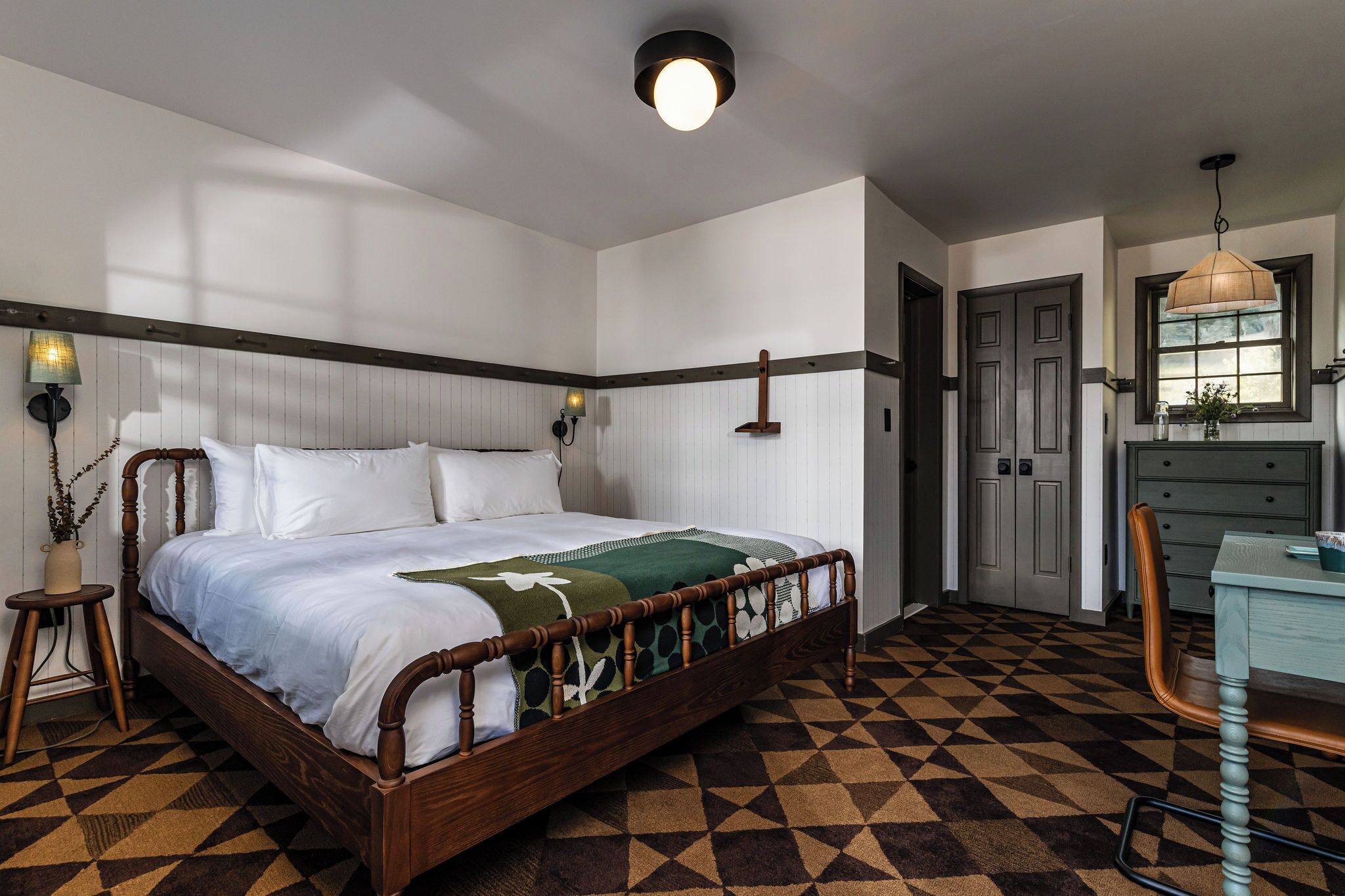 Quarto com cama de madeira, piso xadrez, escrivaninha com cadeira e janela com vista.