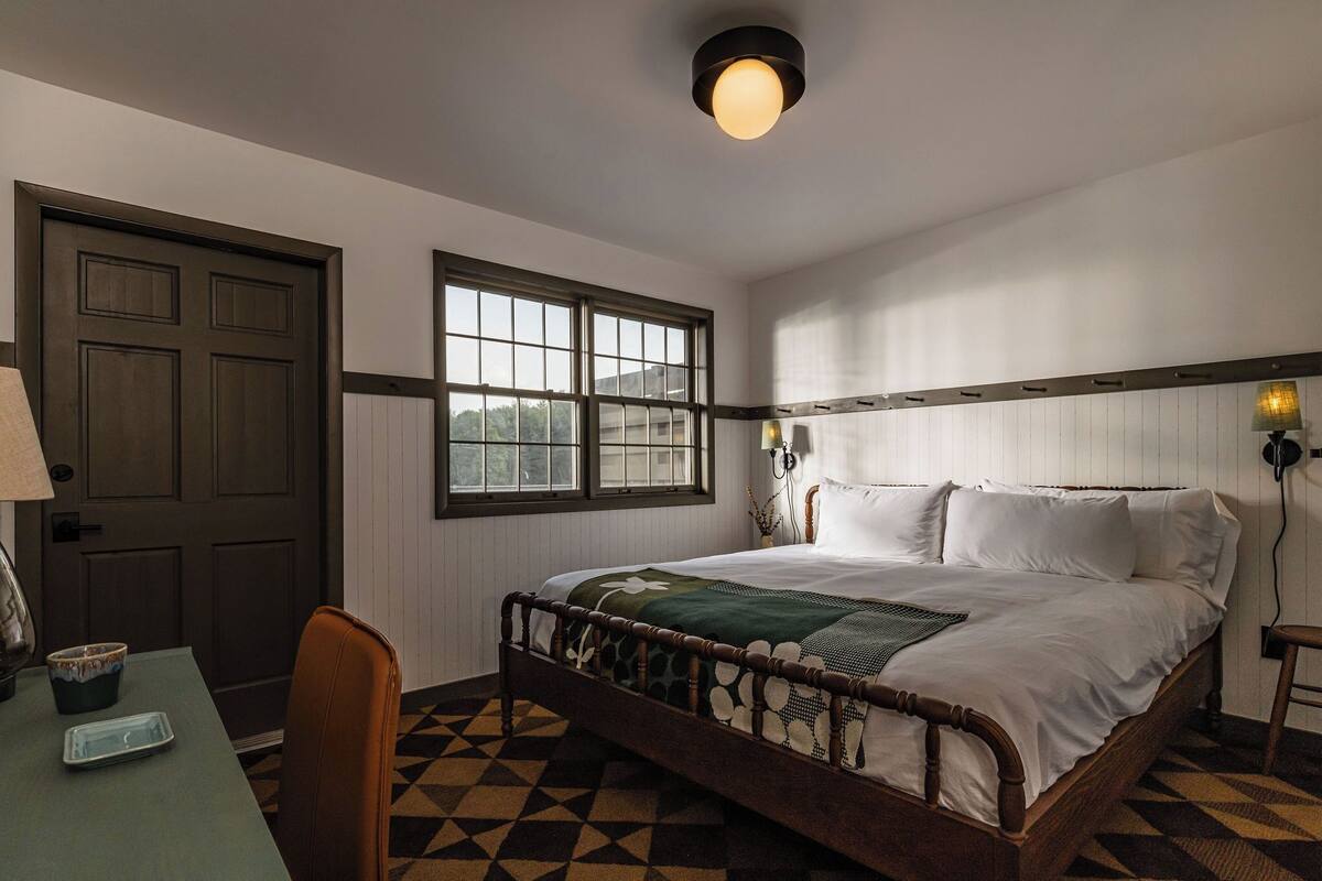 Ein Schlafzimmer mit einem hölzernen Bett, einem Fenster, einem Schreibtisch mit Lampe und einem gemusterten Boden.