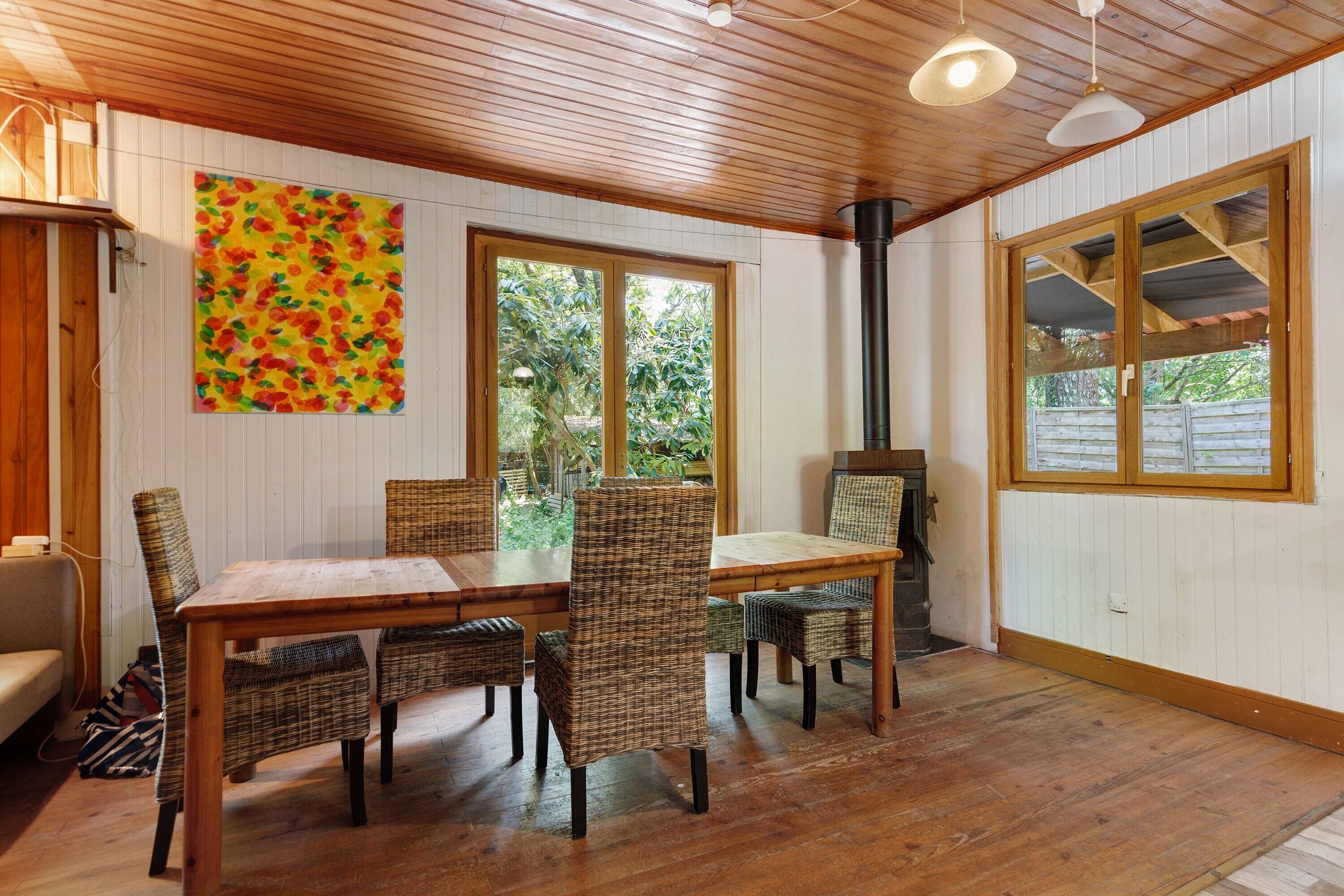 A dining area with a wooden table and wicker chairs, large windows, and a chimney.