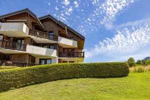 Un immeuble en bois à plusieurs étages, doté de balcons, bordé d’une haie bien entretenue, et surplombé d’un ciel bleu clair.