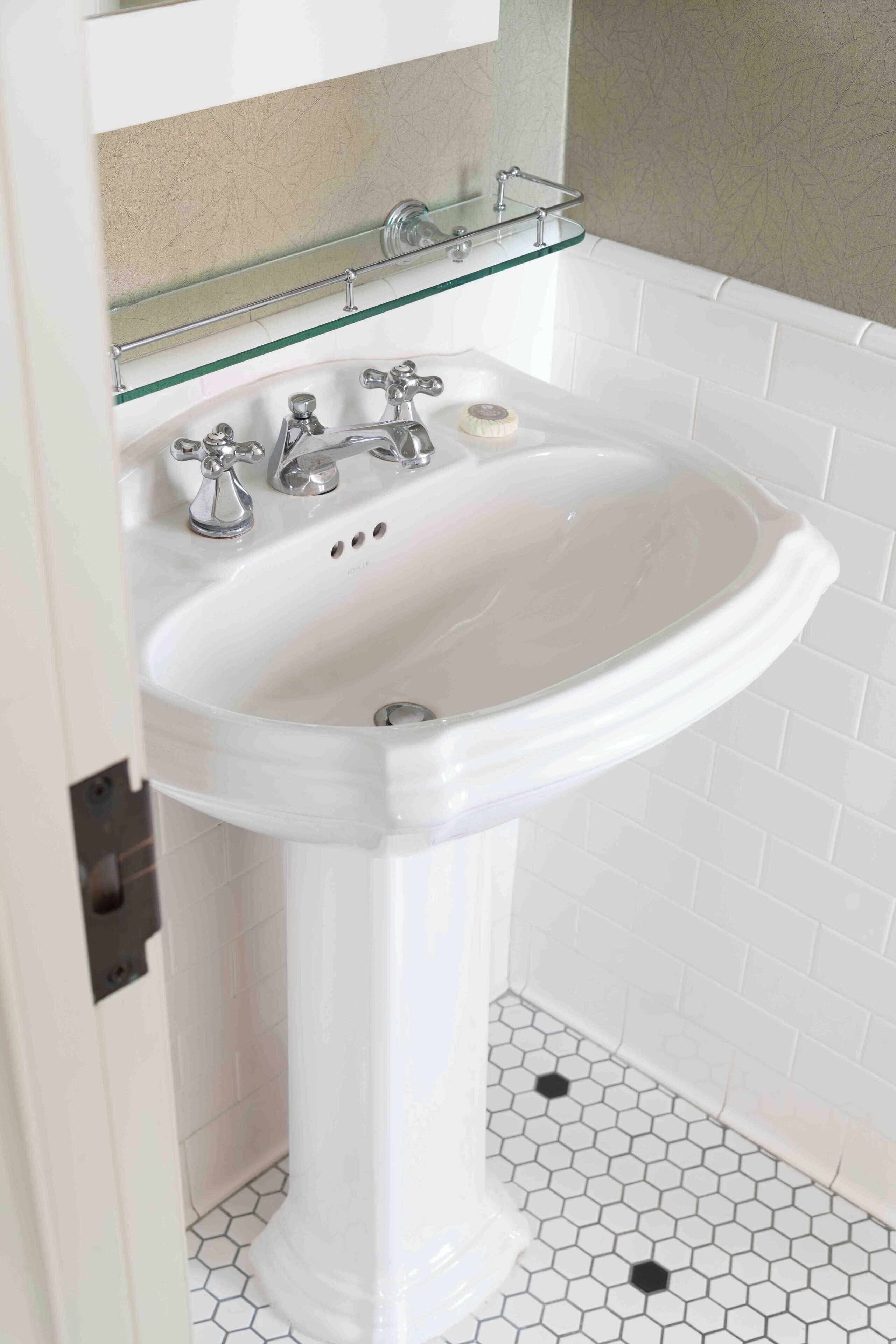 A white pedestal sink with a glass shelf above it, a mirror, and a hexagonal tiled floor.