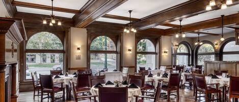 A spacious restaurant with high ceilings, wooden beams, and large arched windows. Tables are set with white tablecloths and red chairs.