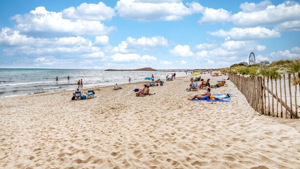 Ein Sandstrand mit badenden und sonnenden Personen, einer Holzumzäunung und einem Riesenrad in der Ferne.