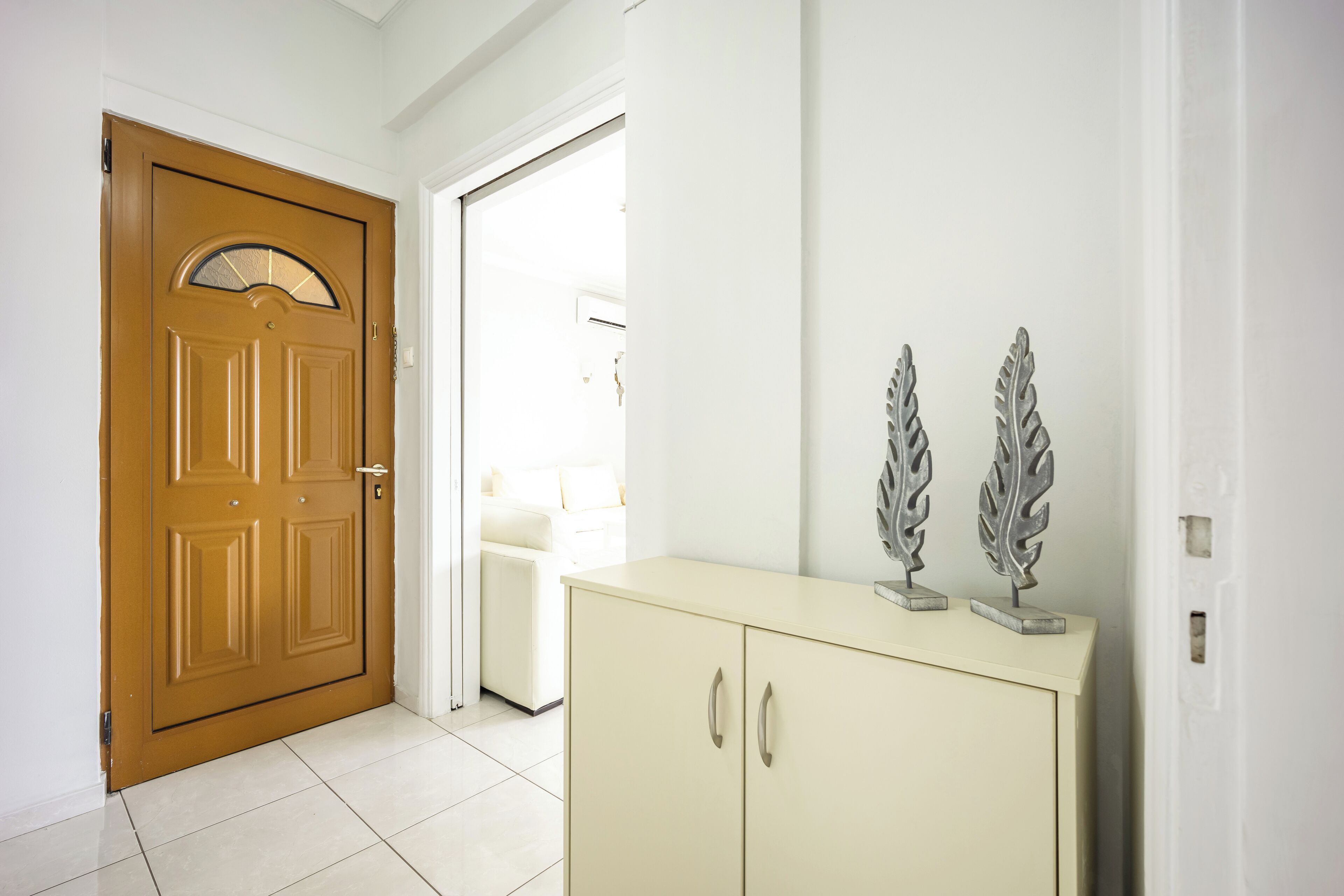 Un couloir avec une porte en bois, un meuble de couleur crème orné de motifs de feuilles métalliques décoratives, et une chaise blanche à l’arrière-plan.