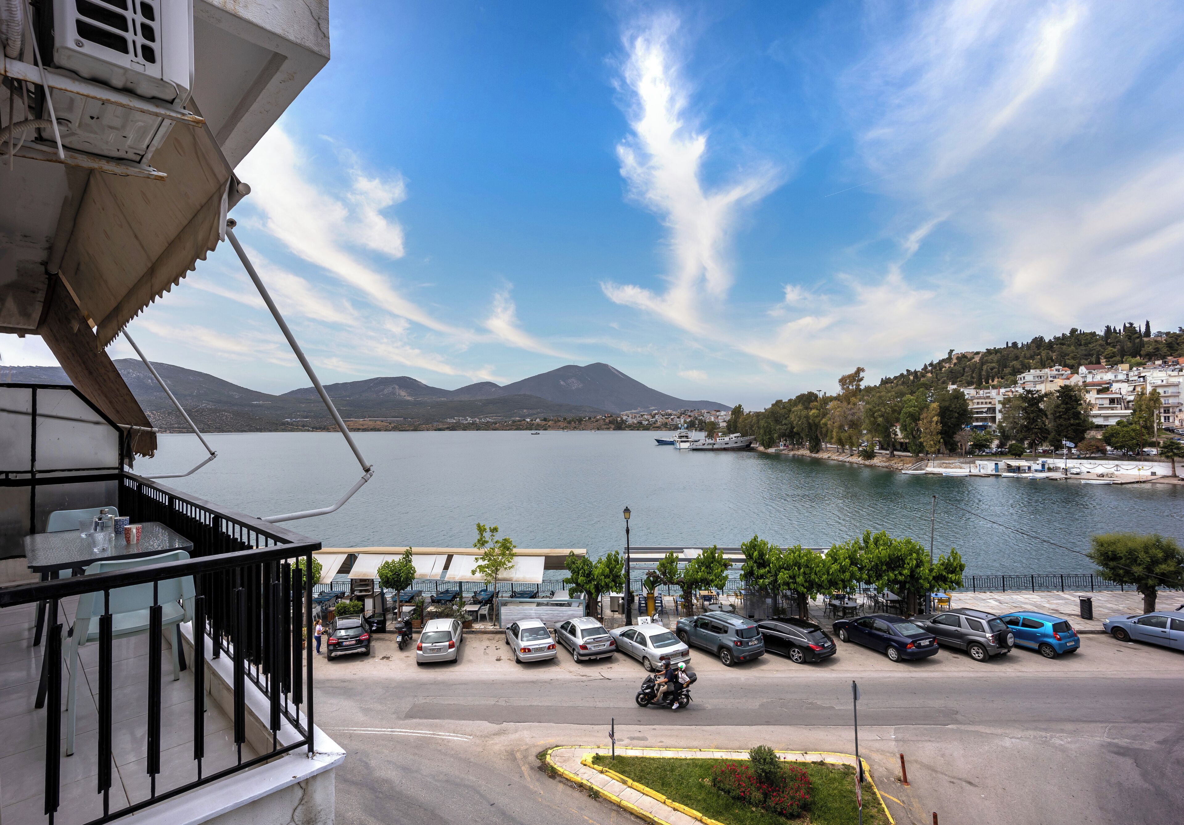 Vista desde un balcón sobre un estacionamiento con coches, una masa de agua y un paisaje montañoso a lo lejos.