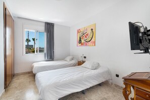 A hotel room with two beds, a TV, a wooden dresser, and a window with a view of palm trees.