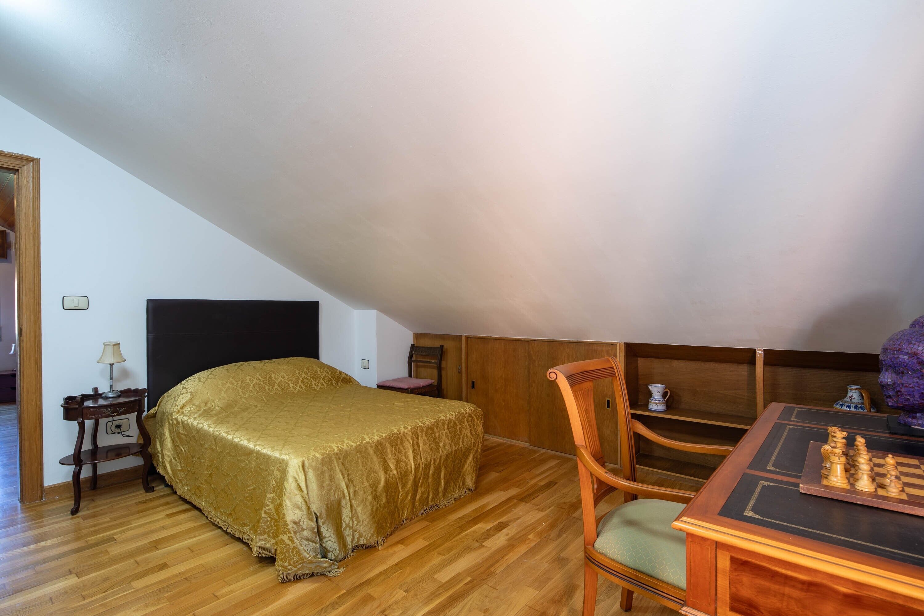 A room with a bed, a wooden table with chess, a chair, a lamp, and a small table with a cup.