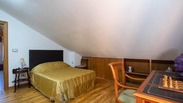 A room with a bed, a wooden table with chess, a chair, a lamp, and a small table with a cup.
