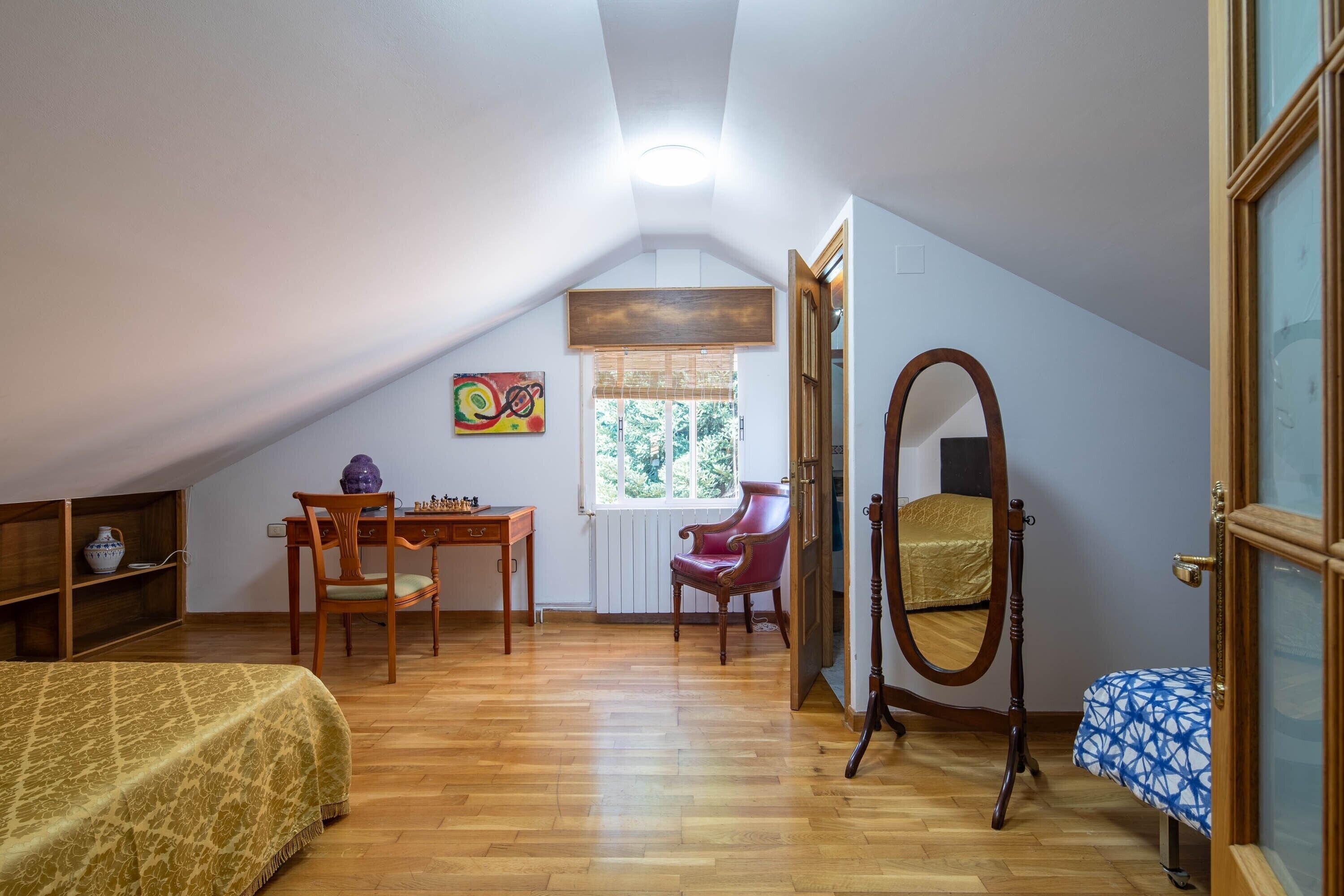 Habitació amb sostre inclinat, escriptori de fusta amb cadira, llit amb llençol estampat, mirall i finestra amb vista a arbres.