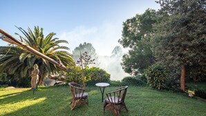 A garden with two wicker chairs and a small round table, surrounded by lush greenery and a statue. - Holiday Home 'O Axouxere' with Mountain View, Private Terrace and Wi-Fi (Feira do Tres)