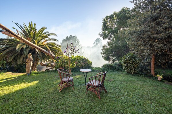 A garden with two wicker chairs and a small round table, surrounded by lush greenery and a statue.