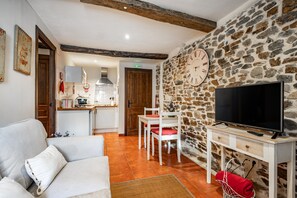 Uma sala de estar com sofá, televisão, parede de pedra e cozinha ao fundo.