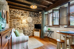 A cozy room with a stone wall, wooden ceiling, a sofa, a television, a small table, and a window with curtains.