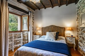 A bedroom with a stone wall, wooden beams, a bed with a blue blanket, two bedside tables with lamps, and a window with curtains.