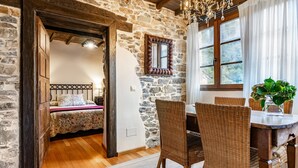 Ein rustikaler Raum mit einer Steinwand, Holzbalkendecke, einem Bett mit Blumenmuster, Korbstühlen, einem Holztisch und einem Fenster mit Vorhängen.