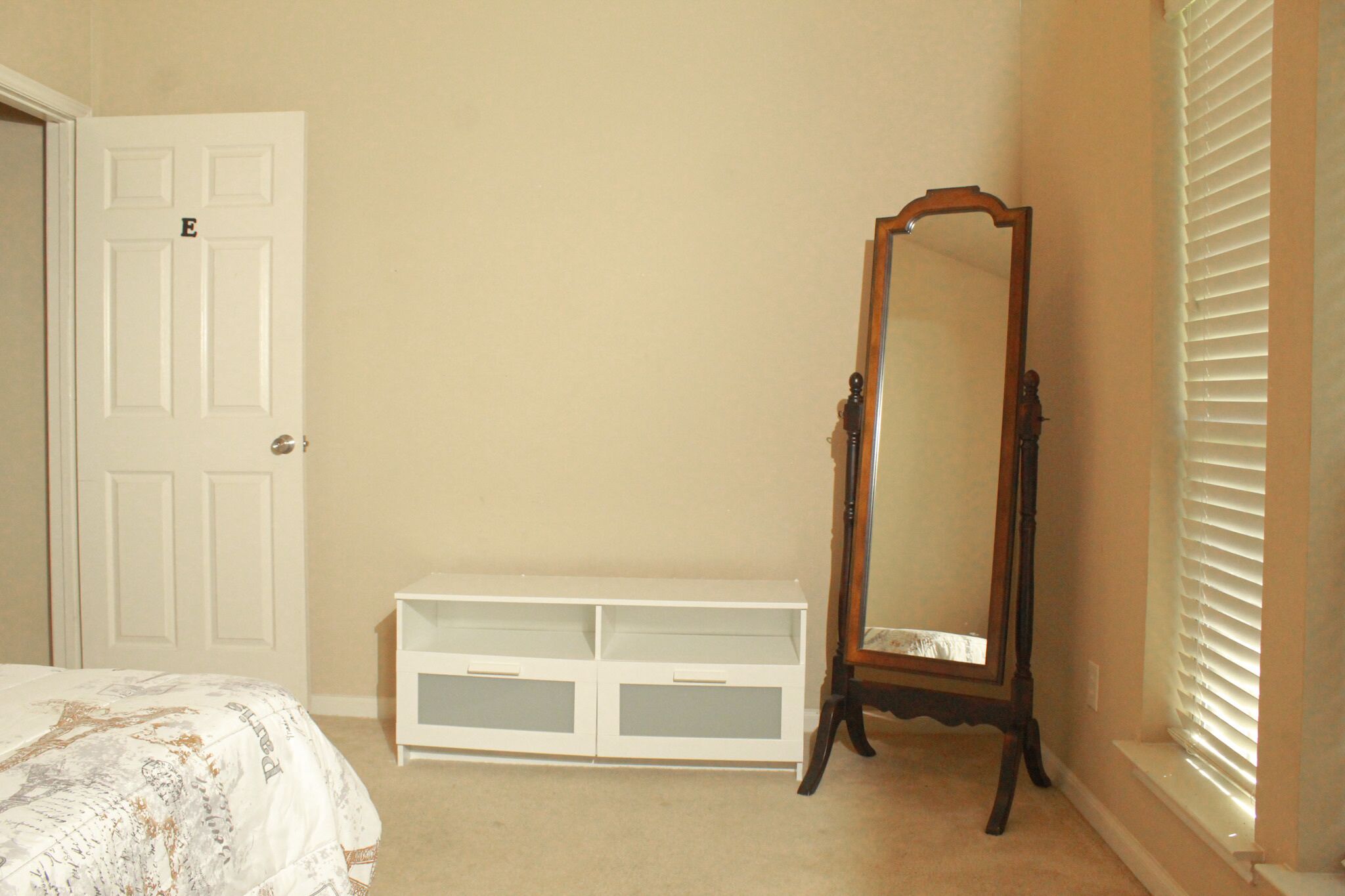 Quarto com uma cama, uma cômoda branca, um espelho de corpo inteiro e uma porta marcada com um 'E'.