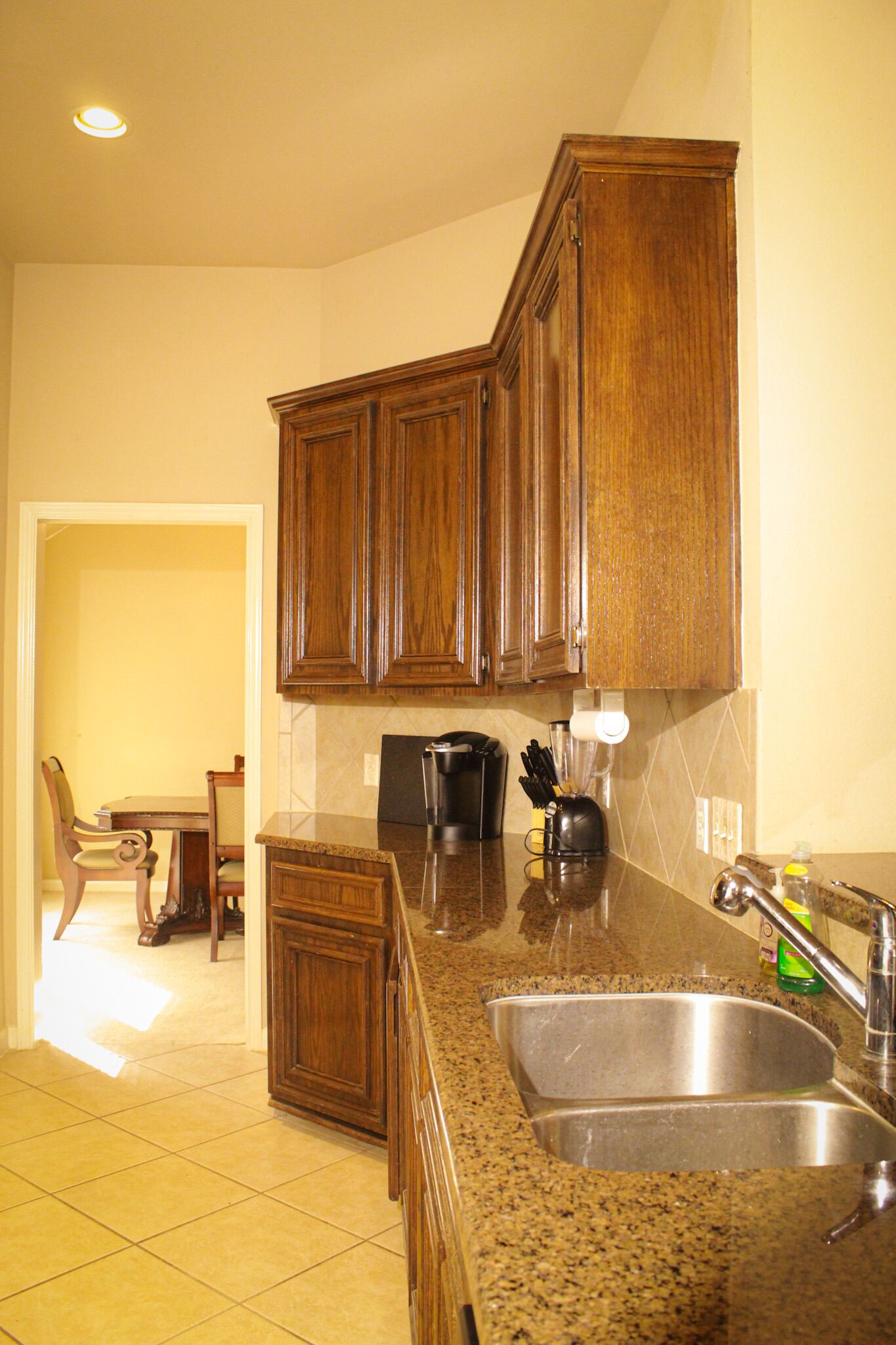 A kitchen with wooden cabinets, a granite countertop, a stainless steel sink, and a dining area with a table and chairs.
