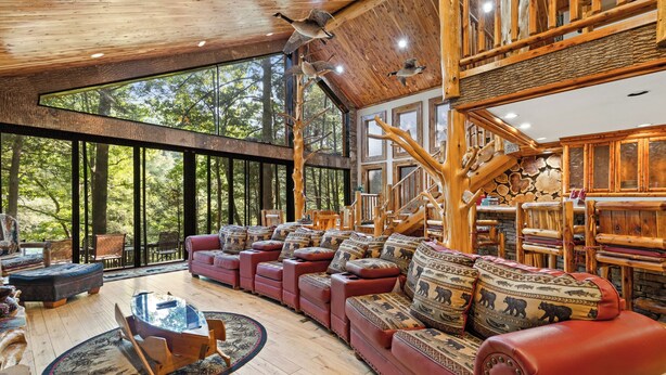 A spacious living area with a large window, wooden furniture, and a fireplace. - Riverside Luxury: Hot Tub, Fire Pit & Fishing Dock! Game Room, Fireplace! (Ellijay)