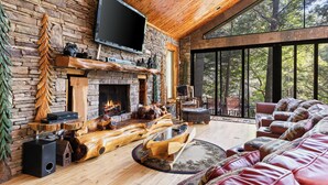 A cozy living room with a stone fireplace, wooden furniture, and a large window overlooking a forest. - Riverside Luxury: Hot Tub, Fire Pit & Fishing Dock! Game Room, Fireplace! (Ellijay)