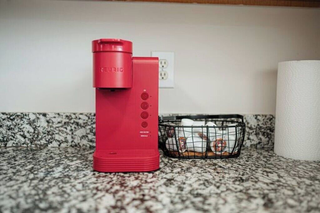 A red electric kettle on a granite countertop next to a black wire basket containing a white cloth and a red object.