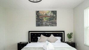 Una cama bien hecha con cubrecama y almohadas blancas, cabecera negra y un cuadro de un puente sobre el agua en la pared.