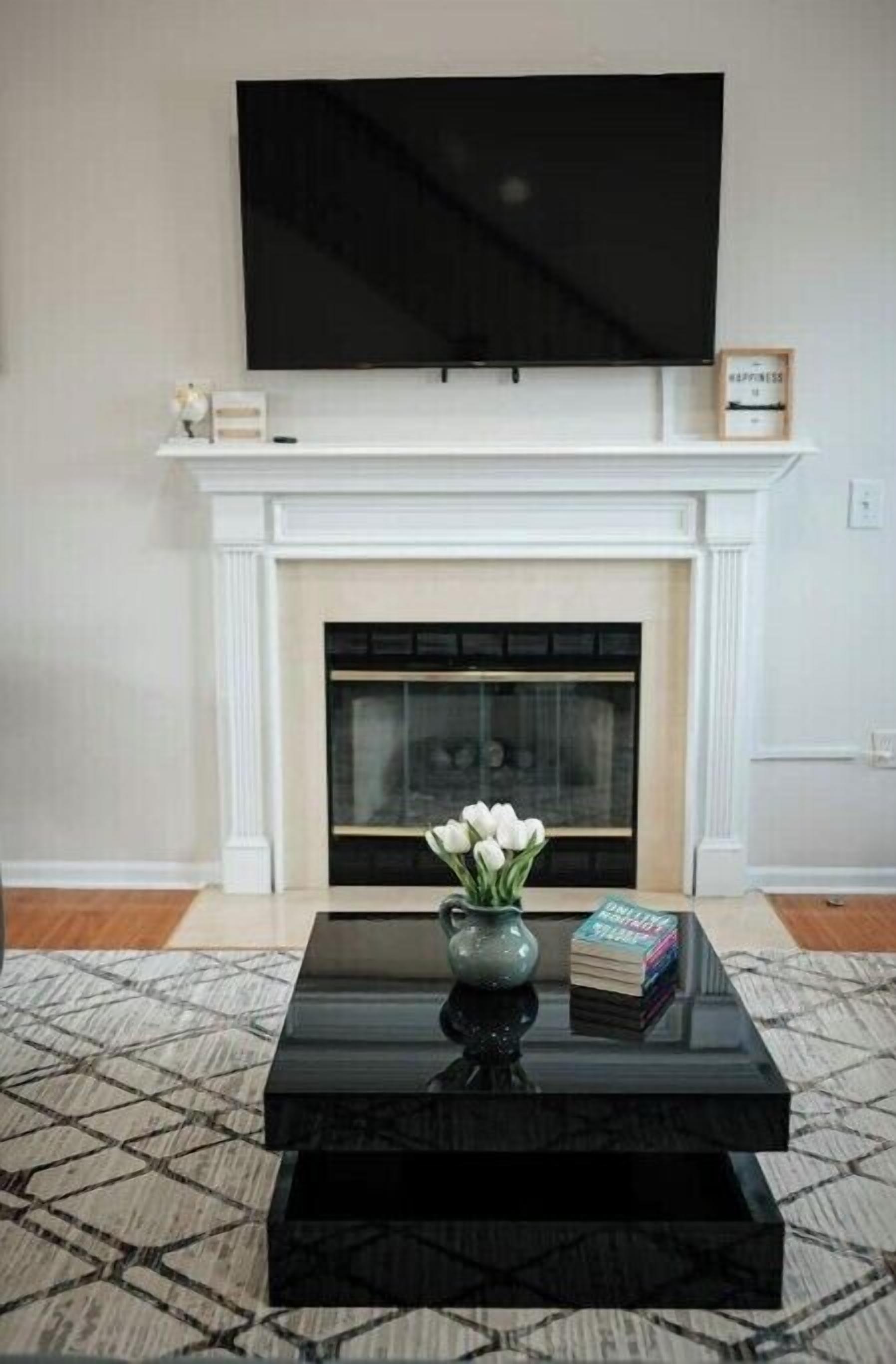 A living room with a fireplace, a flat-screen TV mounted above it, a black coffee table with a vase of white flowers, and a stack of books.
