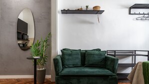 Ein Zimmer mit einem grünen Samtsofa, einem runden Spiegel, einer Topfpflanze und einem Regal mit Dekoration.