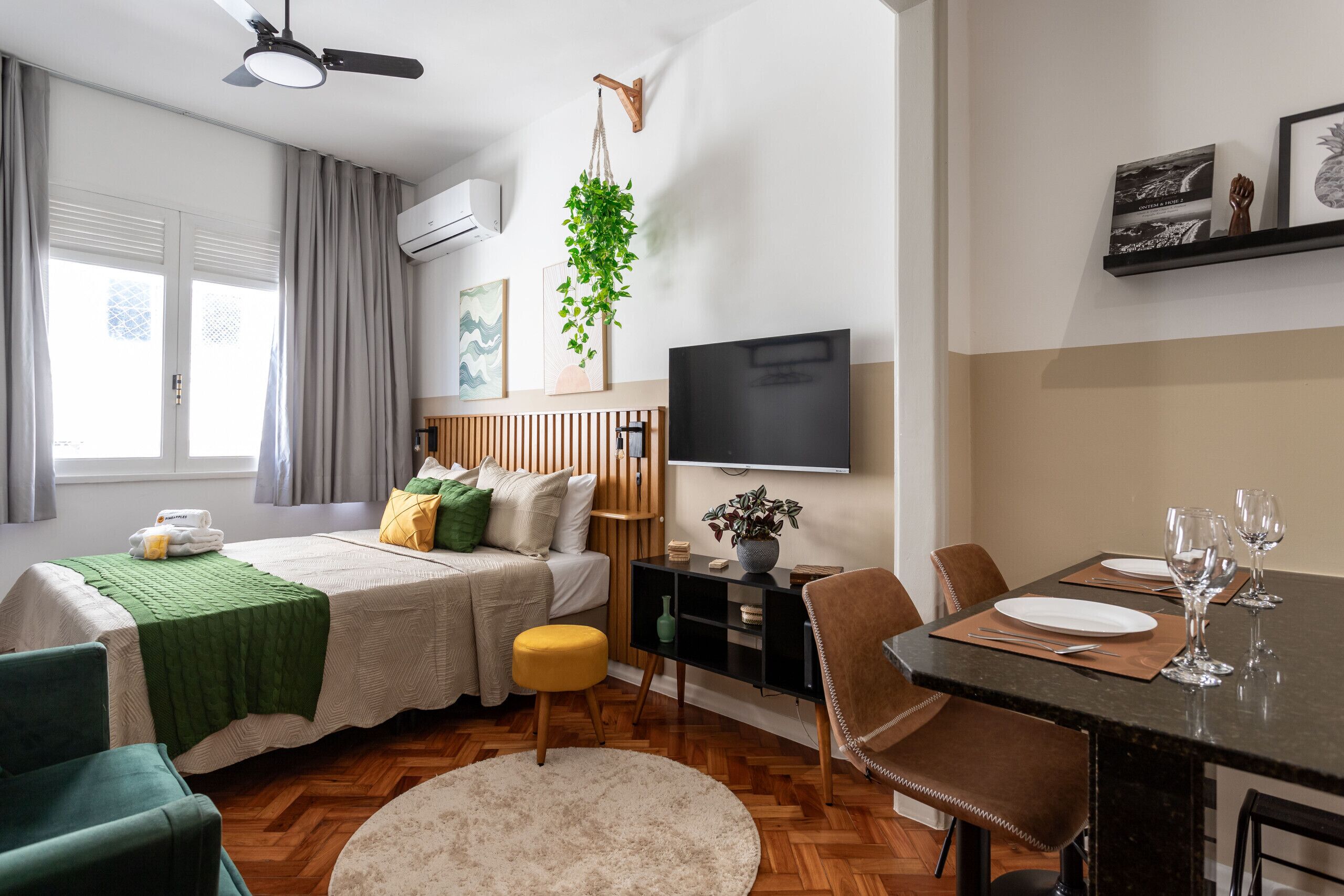 Een moderne, goed verlichte kamer met een bed, een eettafel voor twee personen, een televisie en een hangplant.