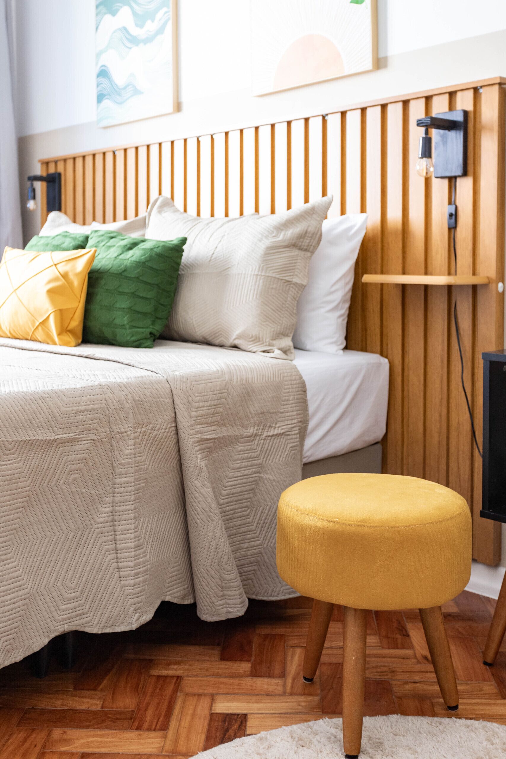 Une chambre à coucher bien rangée, avec un lit, une tête de lit en bois, une table de chevet et un pouf jaune.
