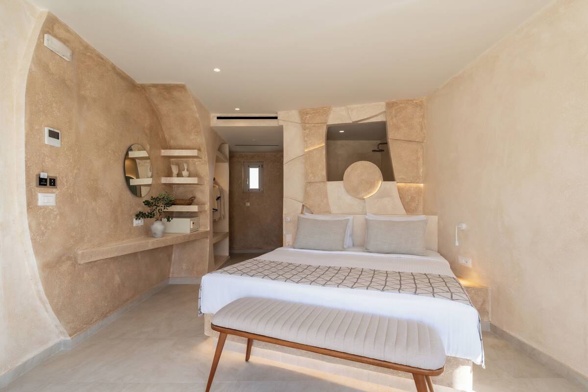 A bedroom with a large bed, a bench, a wall-mounted shelf with decorative items, and a door leading to another room.