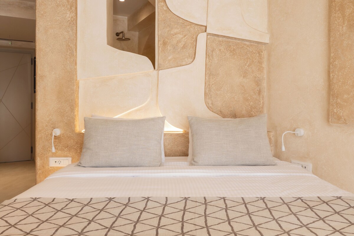 A bedroom with a bed, two pillows, a nightstand, a wall-mounted lamp, and a mirror.
