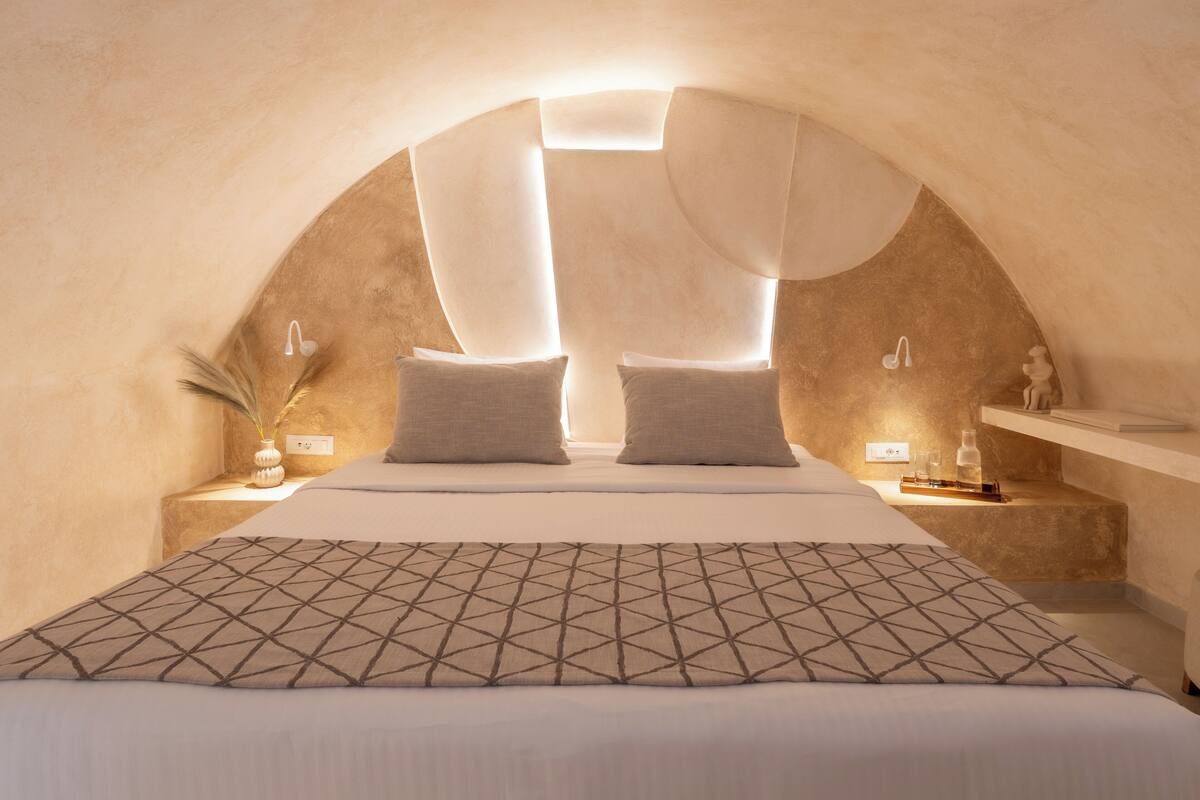 A bedroom with a vaulted ceiling, a bed with patterned bedding, bedside tables with lamps, and a shelf with decorative items.