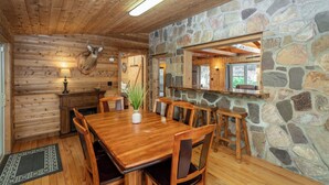 A wooden interior with a stone wall, a wooden dining table with chairs, a mounted deer head, and a stone fireplace.