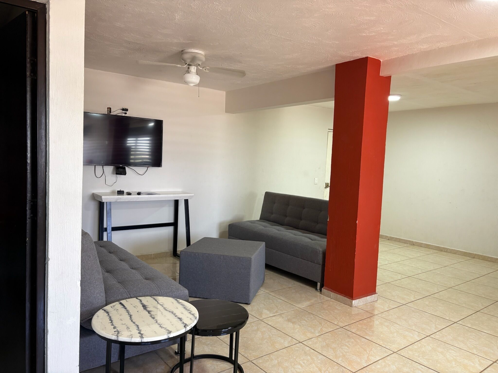 Un salon avec un canapé gris, une petite table ronde, un téléviseur fixé au mur et un ventilateur de plafond.