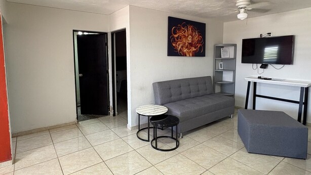 A living room with a grey sofa, a small round table, a grey ottoman, a flat-screen TV mounted on a white stand, and a ceiling fan. - Lomas Altas Tonala Céntrico 8 (Tonalá)