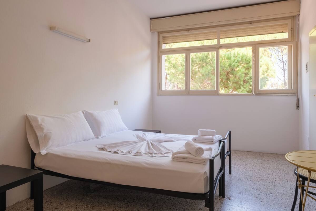 A clean, minimalist bedroom with a bed, pillows, a bedside table, a small table, and a window with a view of trees.