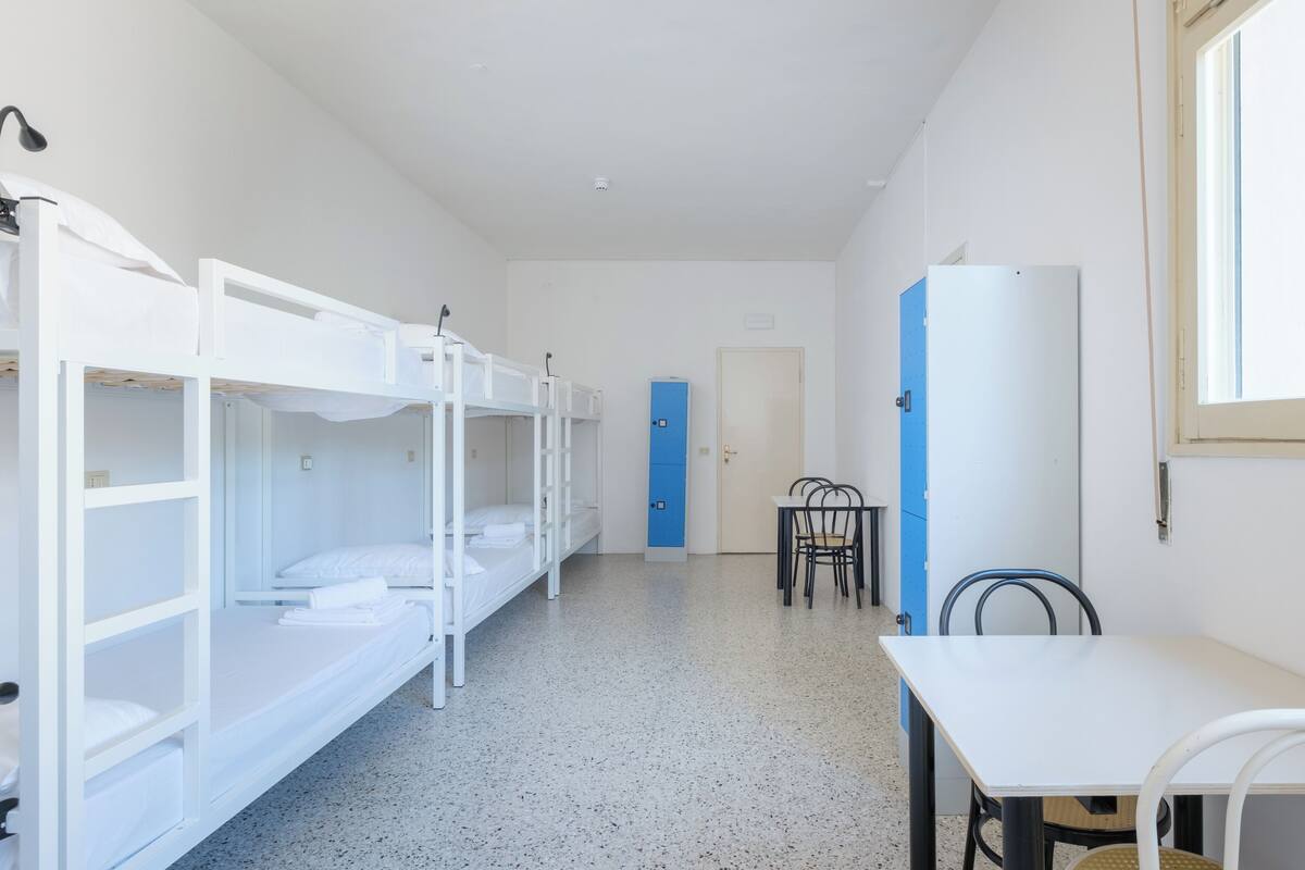 A dormitory room with bunk beds, a small table with chairs, a blue door, and a white refrigerator.