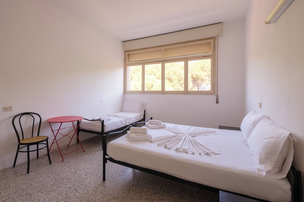 A small, clean room with a single bed, a small table, a chair, and a window with blinds.