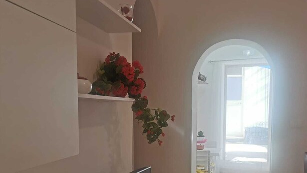 Een moderne woonkamer met een flatscreen televisie bevestigd aan een wit kastje, een klein plankje met decoratieve objecten, en een gang naar een lichte kamer met een raam.