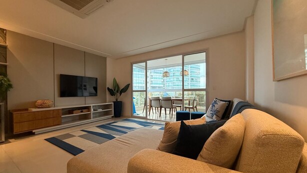 A modern living room with a large sofa, a flat-screen TV, a dining area with a table and chairs, and a view of a building through large windows. - Enseada Azul - Beachfront, Luxury (Guarapari)