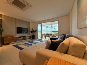 A modern living room with a large sofa, a flat-screen TV, a dining area with a table and chairs, and a view of a building through large windows. - Enseada Azul - Beachfront, Luxury (Guarapari)