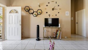 Een woonkamer met een witte deur, een wandklok, een flatscreen televisie, een zwarte speaker en een kleine potplant.