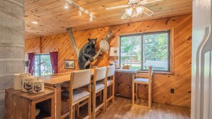 A wooden interior with a bear sculpture on a tree branch, a dining table with chairs, and a window with a view of the outdoors.