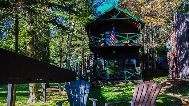 A cabin with a green roof, surrounded by trees and two Adirondack chairs in the foreground.