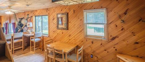 A wooden cabin interior with a dining area, a fireplace, and a large window.