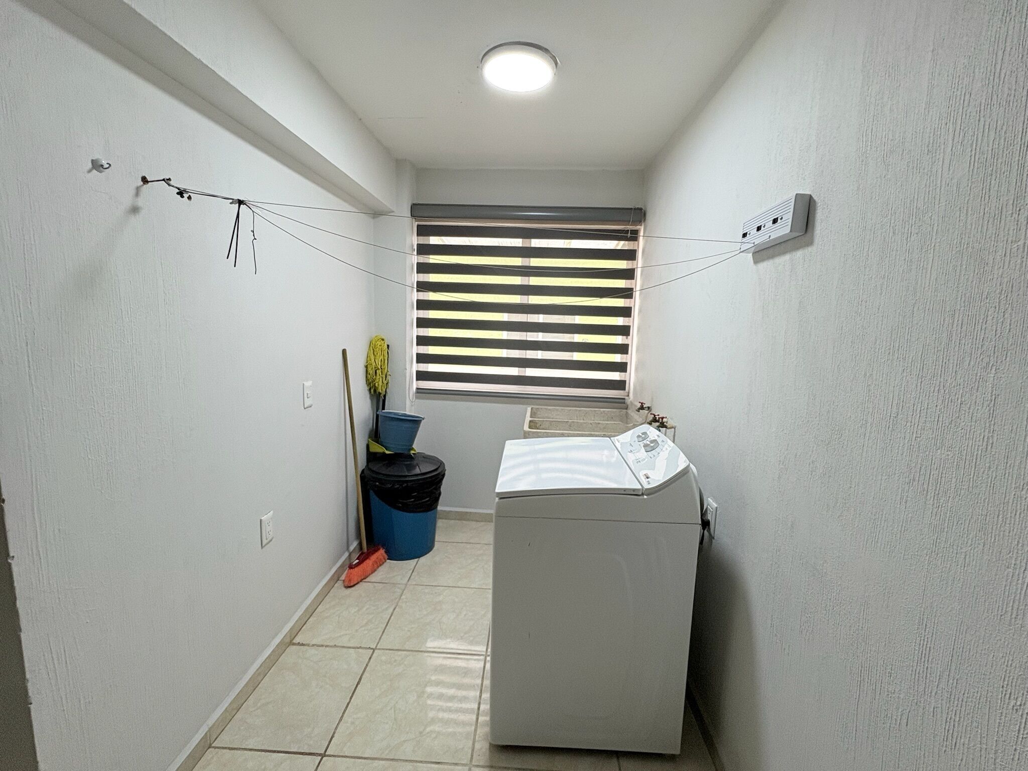 A laundry room with a washing machine, a window with blinds, a mop, and a broom.