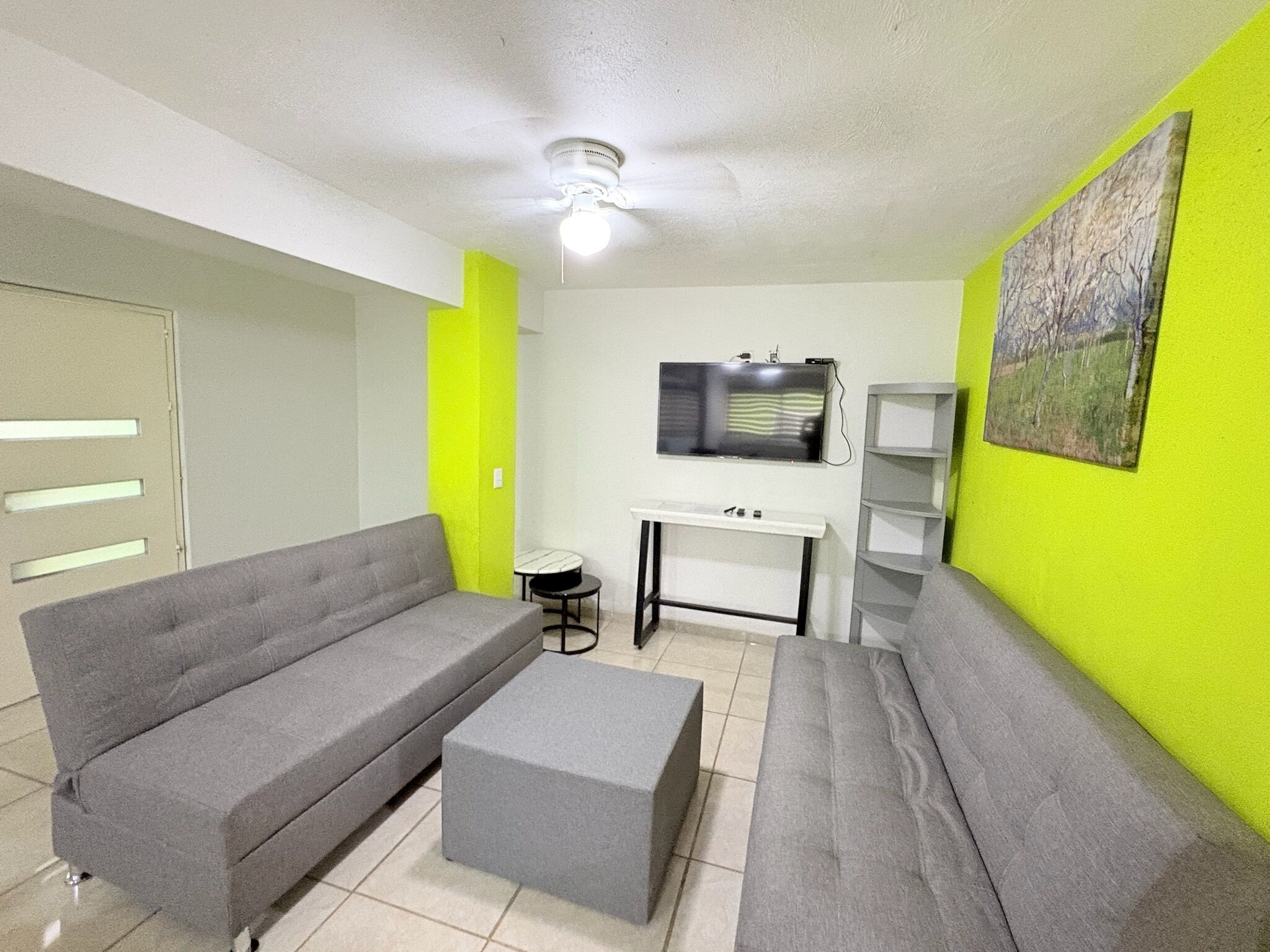 Un salon avec un canapé gris, un pouf gris, un téléviseur posé sur un meuble blanc et un mur vert.